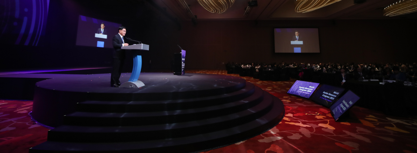 Man in suit at podium on stage with audience and screen showing speaker at World HR Congress 2024.